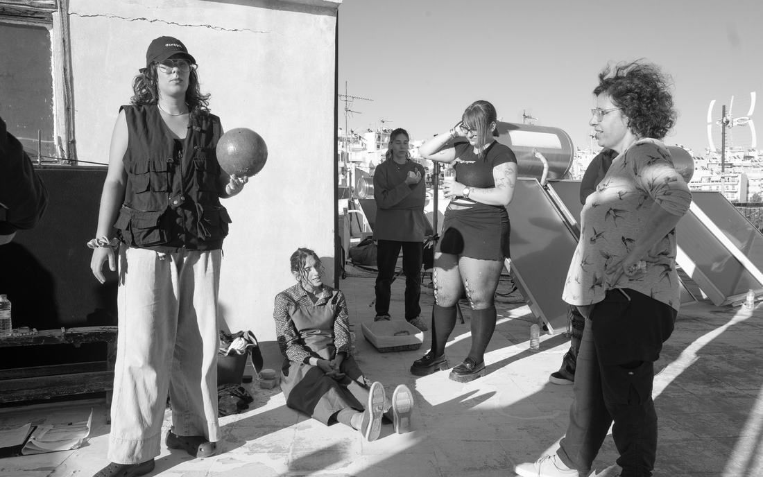 Black-and-white still on a sunny Athens rooftop. Five women stand and sit among solar water heaters; one holds a ball.