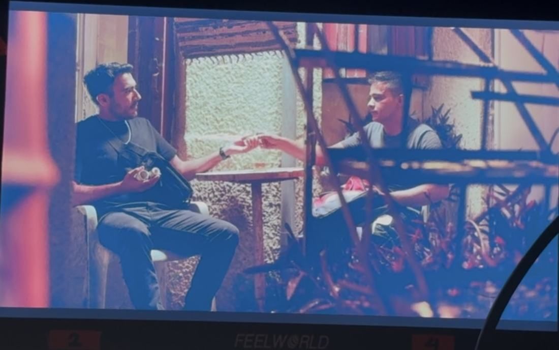 A field monitor shows two men at a small table in warm light, one handing something to the other across it.