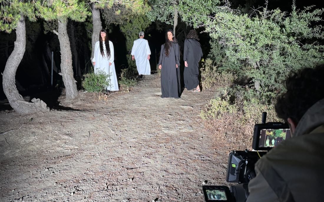 Night shoot in a pine forest. Four performers in long white and black robes stand on a dirt path while a camera operator frames them on a monitor.