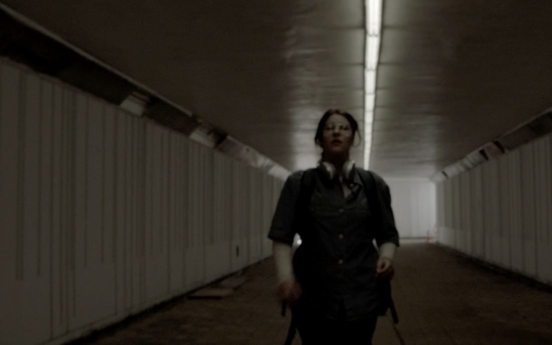 A woman with headphones around her neck walks toward the camera through a long, dim pedestrian underpass under a strip of fluorescent light.