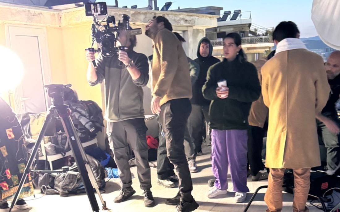 Behind the scenes on a rooftop at golden hour. A camera operator lifts a rig while cast and crew wait between takes.