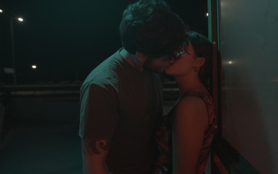A couple kisses against a glass wall at night, lit in deep teal and red, with streetlights blurred behind them.